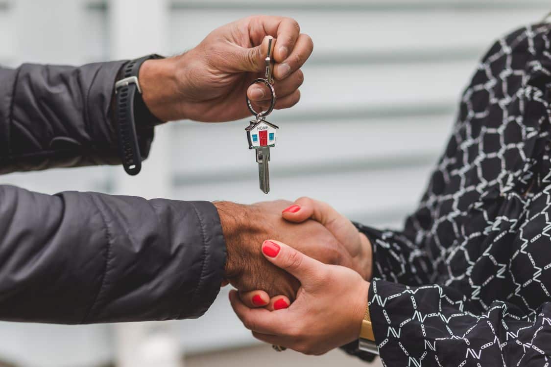 A person's hand holding a set of new house keys, with the blurred background of a beautiful, modern home on a sunny day.